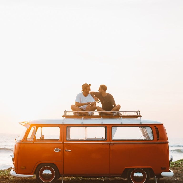 Zwei Personen sitzen auf einem orangefarbenen VW-Bus am Strand bei Sonnenuntergang.
