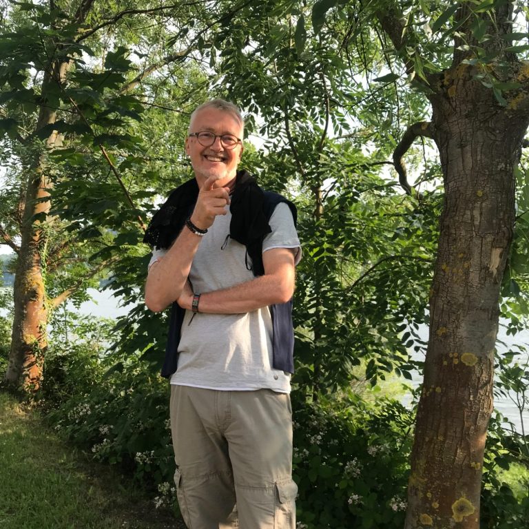 Frank steht lächelnd im Freien vor Bäumen und Sträuchern.