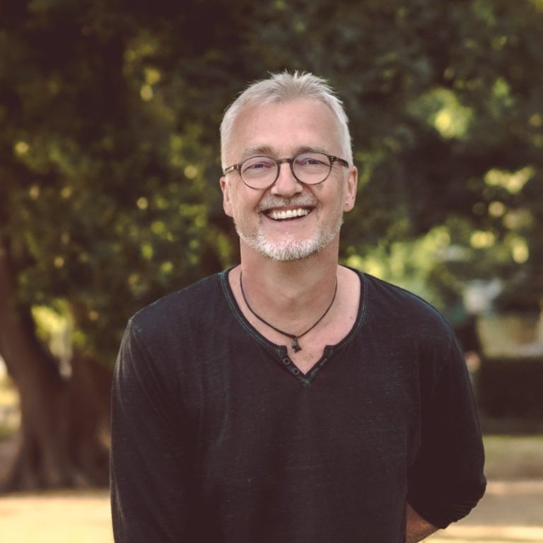 Lächelnder Frank mit seinen grauen Haaren, im Freien vor Bäumen stehend.