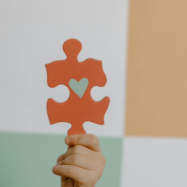 Eine Hand hält ein rotes Puzzlestück mit einem grünen Herz auf einem bunten Hintergrund.