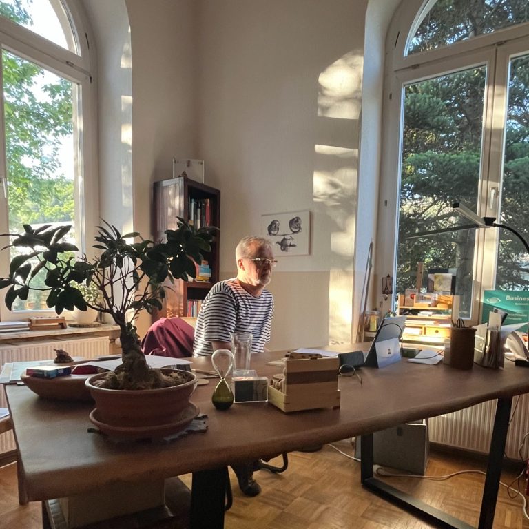 Frank sitzt an einem Tisch mit Pflanzen und Fensterlicht in einem hellen Raum.